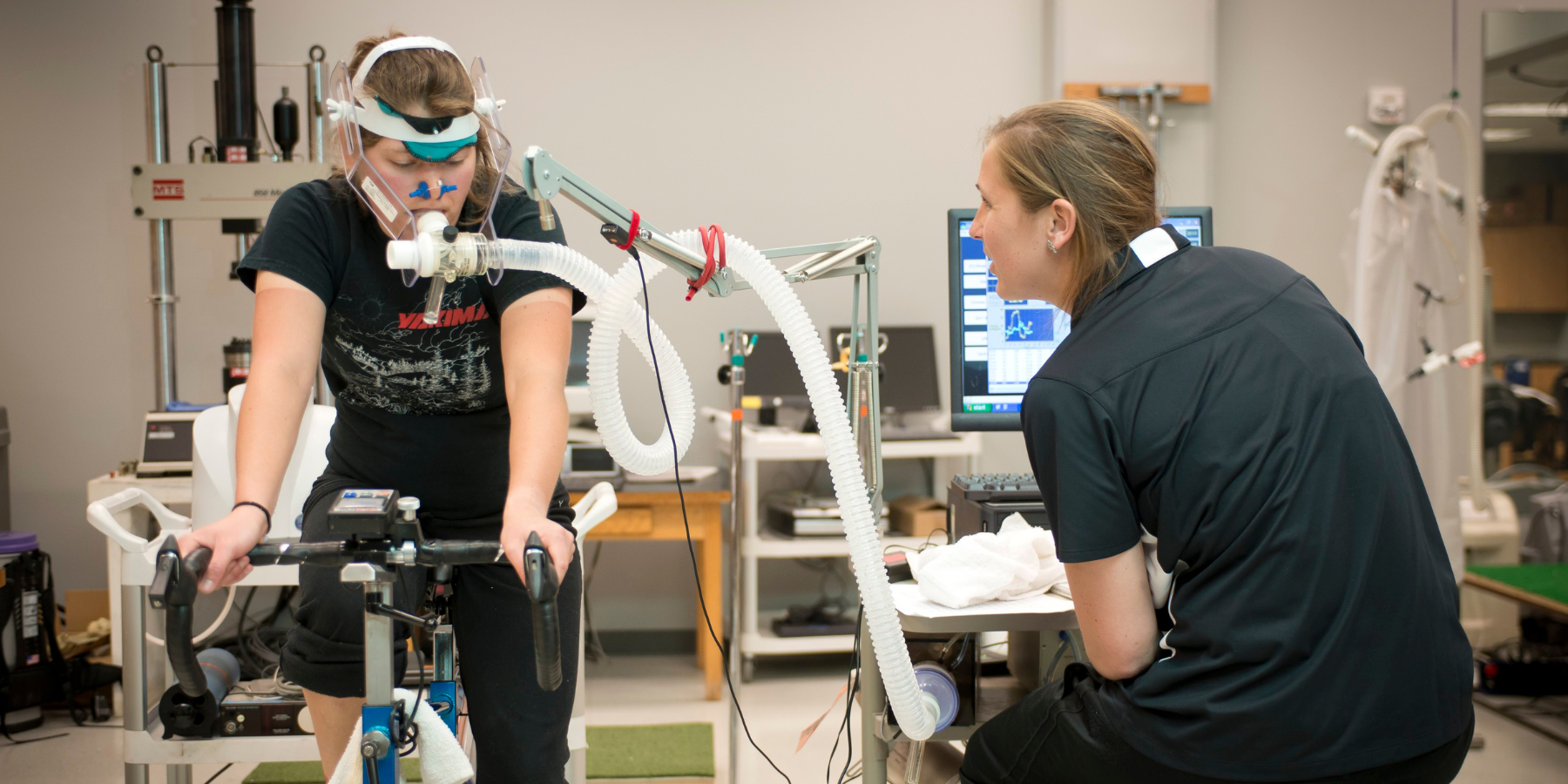 Photo of two students working in a lab, measuring health stats while someone is riding a stationary bike.