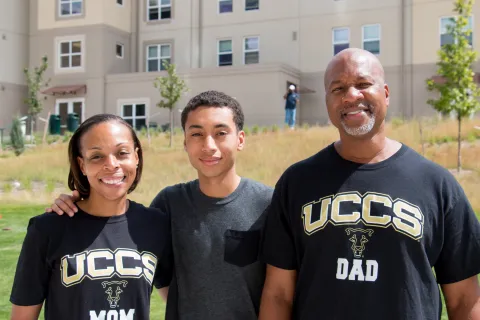 A student standing in front of the Alpine Village apartments with his mom and dad.