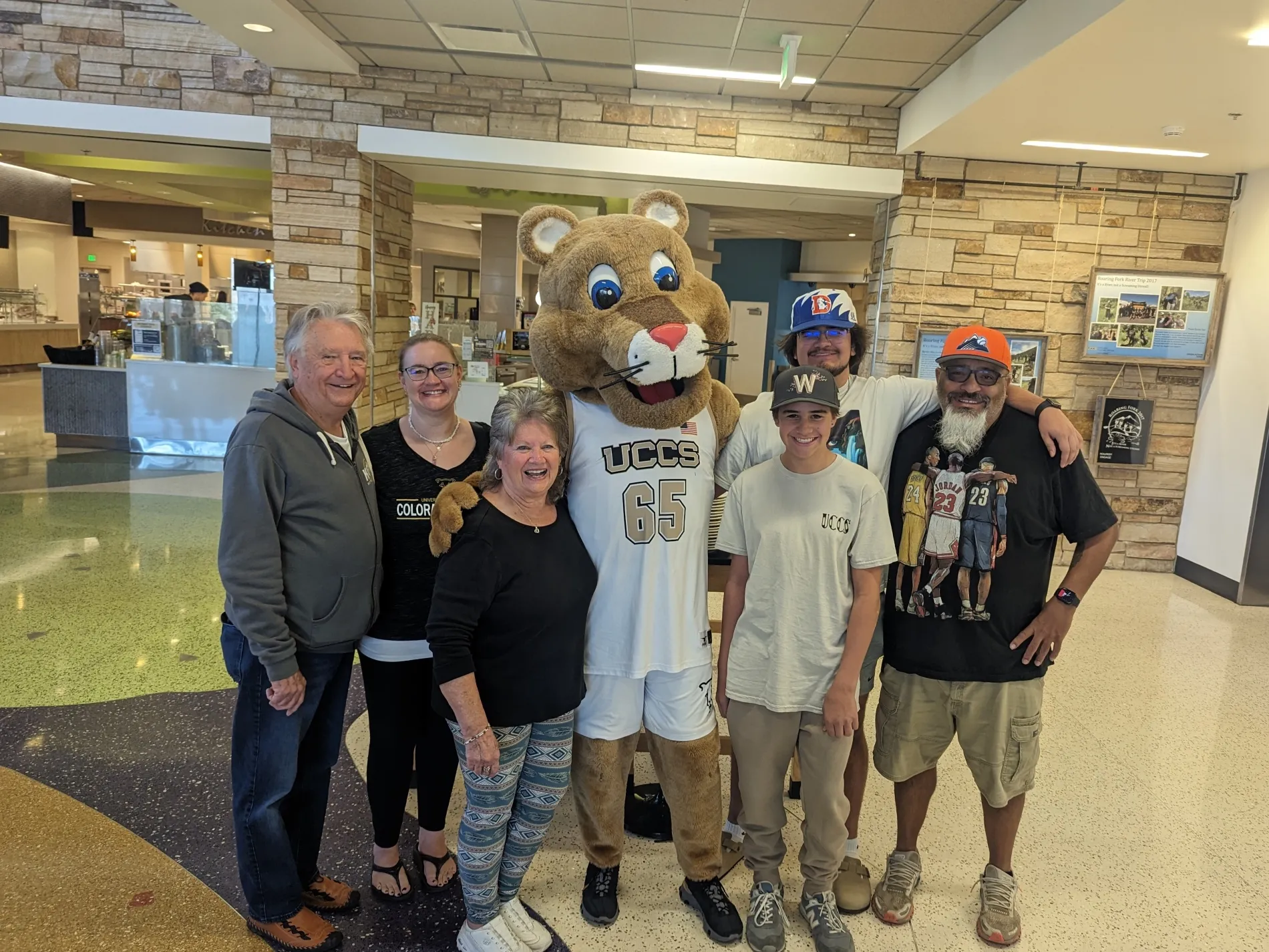 A family of six posing for a photo with mascot, Clyde at the UCCS Roaring Fork Dining Hall.